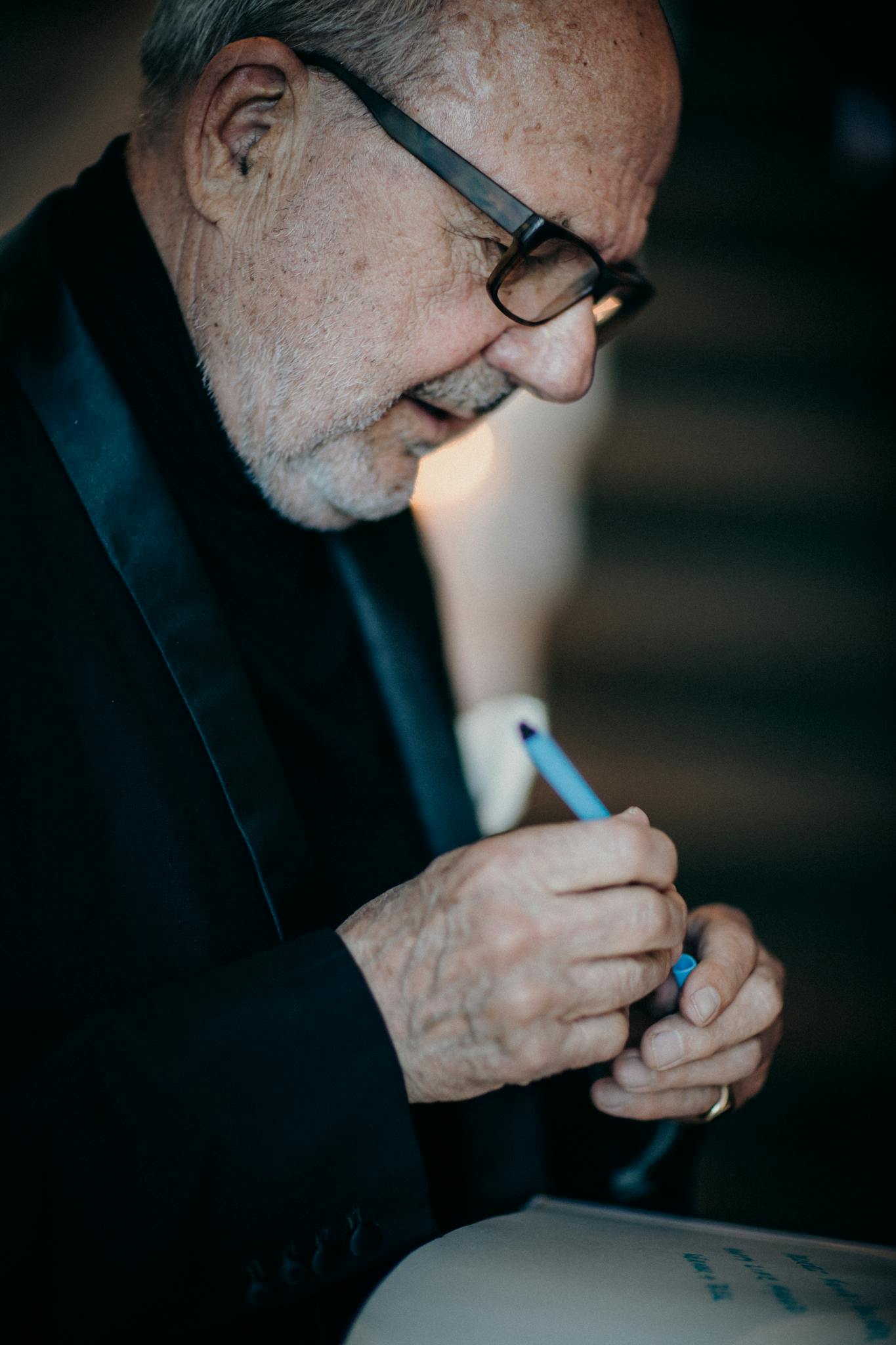 Home 3 Elderly Caucasian man in glasses attentively writes with a blue pen in an elegant indoor setting.